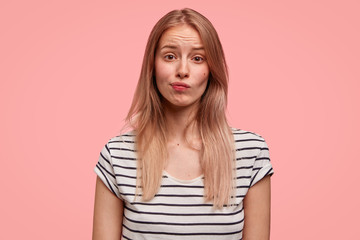 Portrait of displeased female with gloomy expression, frowns face, has healthy skin, wears striped casual t shirt, looks embarrassed at camera, isolated on pink background. Negative facial expressions