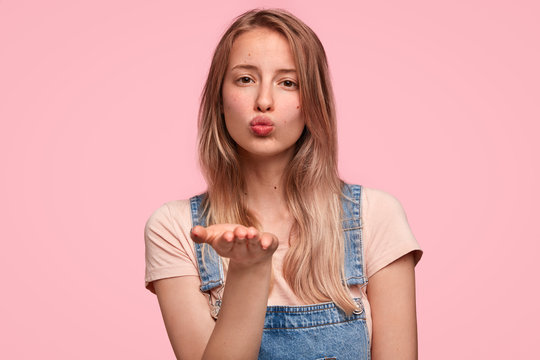 Close up portrait of attractive young Caucasian female blows air kiss at camera as flirts with her boyfriend, has relaxed facial expression, holds palm near mouth, sends love to surrounding.