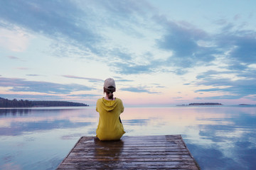  A girl sits on a wooden pier against a background of evening sunset over a lake at full  calm water