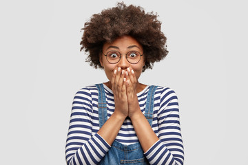 Photo of amazed excited joyful female with Afro hairstyle, covers mouth and exclaims with...