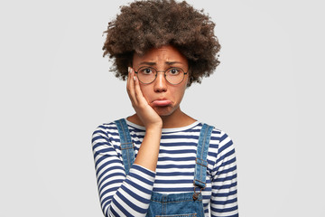 Dull doleful young African American female has sorrowful expression, purses lips and keeps hand on cheek, being abused by her husband, finds out about betrayal, isolated over white background