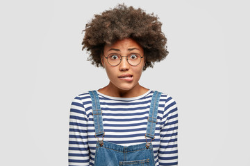 Embarrassed beautiful African American female bites lower lip nervously, raises eyebrows and has puzzled expression, dressed in casual clothing, isolated over white background. Emotions and ethnicity