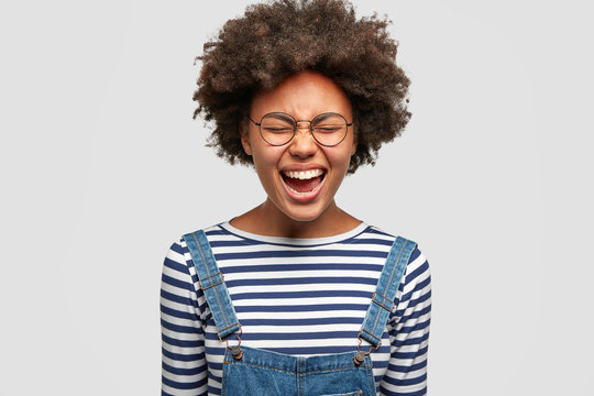 Pretty mixed race female model laughs happily, being in high spirit, smiles joyfully, shows white teeth, expresses truthful sincere feeling, keeps eyes shut and mouth opened. Positiveness concept