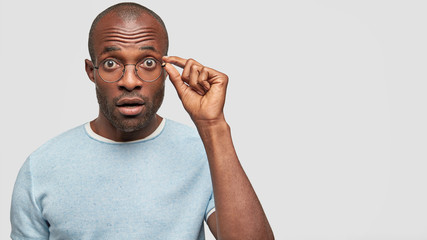 Emotional young African American guy stares through spectacles, has surprised expression, keeps...