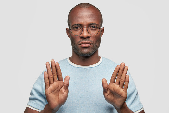 Serious Dark Skinned Male Shows Stop Gesture, Tries To Reject Doing Something, Has Serious Expression, Wears Light Blue T Shirt And Spectacles, Isolated On White Background. Refusal And Body Language