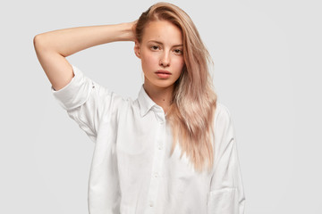 Indoor shot of cute young female with long hair, has healthy pure skin, dressed in loose white shirt, demonstares her natural beauty, poses against studio wall. People, youth, monochrome concept