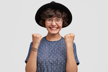 Happy Caucasian cute young female model clenches fists, celebrates her success, smiles broadly, shows white perfect even teeth, dressed in elegant hat and blouse, isolated on white background.