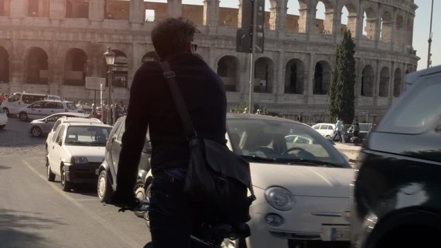Young hipster man riding bike on the road around colosseum in Rome city centre on sunny day slow motion camera car