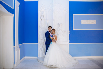 The bride and groom are posing for a photo in a huge blue hall with white columns. The color of the groom's costume is combined with the color of the walls and the bouquet of the bride.