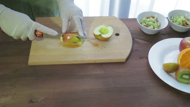 A woman cuts an apple with a knife into slices on a wooden board. In the background is a plate of fruit.