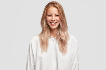 Cheerful beautiful European female with pleasant smile, has white perfect teeth, wears oversized shirt, has straight hair, laughs on funny joke. People, positive emotions and feelings concept