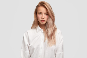 Waist up shot of serious Caucasian female with straight luxurious hair models in studio, dressed casually, poses for salon advertisement, isolated over studio wall. People and lifestyle concept