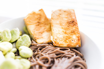 Soba buckwheat noodles Japanese food fried baked golden crispy tofu, green fava beans on hot, cold summer, white bowl macro closeup