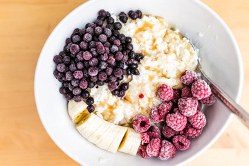 Oatmeal oat grain rice pudding porridge bowl macro closeup with frozen berries, raspberries, blueberries, bananas and maple syrup caramel on wooden table, spoon