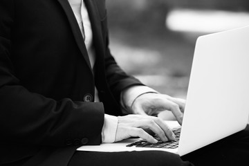 Closeup View of young businessman in formalwear typing on laptop keyboard while sitting at city green grass outdoor. Blurred background.Black and white.