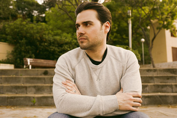 Portrait of Pensive cute young caucasian hipster in informal white jersey sitting at sunny city park.Blurred background