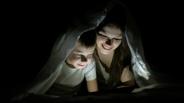 Young woman mom and her son watching a funny film together on tablet under the blanket.