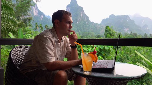 Cinemagraph of Male Traveler Using Laptop with Scenic Limestone Rocks View. Digital Nomad Working on Terace. Freelancer Typing on Computer in Asia. Remote Work in Thailand