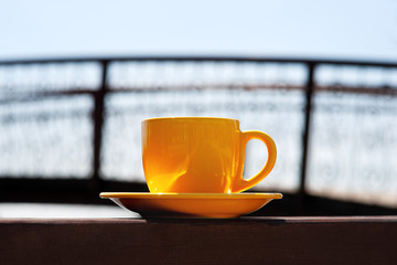 A yellow cup of tea against the background of the bridge and sky. Tea in a yellow cup against the blue sky and railings of the bridge. Romantic rest.