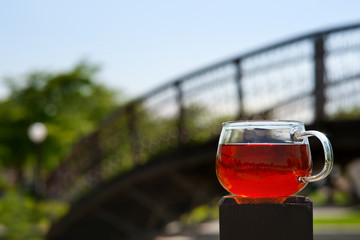 Tea under the summer sun. Transparent cup of tea on the background of the bridge, sky and greenery.