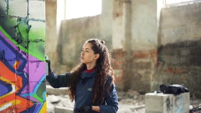 Slow motion of graffiti artist painting on wall in abandoned building using aerosol spray paint. Attractive girl with curly hair is busy with her work, she is looking at painting.