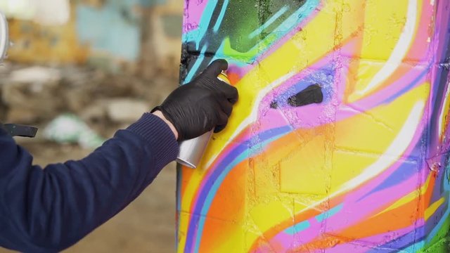 Close-up shot of male hand in leather glove holding spray paint and painting graffiti on old pillar inside abandoned building. Modern art, abstract images and people concept.