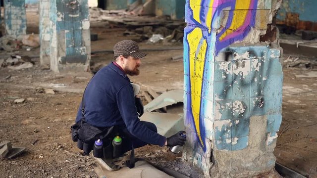 Graffiti artist bearded guy is painting on pillar in abandoned building with aerosol paint spray. Empty industrial building with dirty walls and floor is in background.