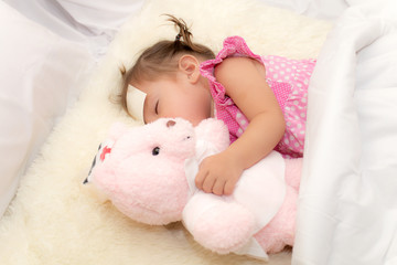 Fototapeta na wymiar Baby drinking water or milk from a small bottle and lying on bed while hugging pink teddy bear in the bedroom because the baby having fever and attach cooling gel pad on her forehead for relief fever.