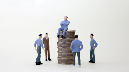 A miniature man sitting on a pile of coins next to a four miniature men standing.