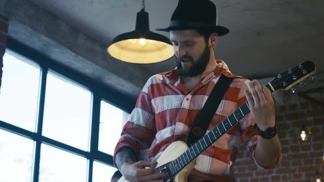 From below shot of bearded man in hat and checkered shirt playing guitar and singing in loft studio