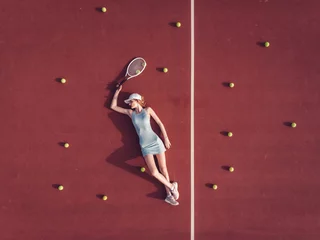 Zelfklevend Fotobehang Overhead top view of young Caucasian teen model wearing fashionable tennis dress, lying on tennis hardcourt with a lot of balls, summer sunny day outdoors. Fashion portrait shoot © supamotion