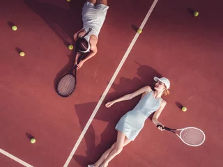 Foto op Canvas Overhead top view of two young Caucasian teen models wearing fashionable tennis dresses, lying on tennis hardcourt with a lot of balls, summer sunny day outdoors. Fashion portrait shoot © supamotion