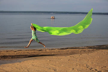 Młoda dziewczynka skacze przez wodę z długą zieloną chustą. - obrazy, fototapety, plakaty