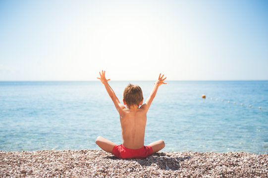 happy little kid sitting on sea shore beach raised his hands to summer sun