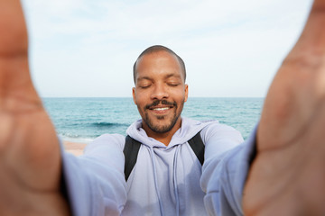 Handsome young African-American hipster man making smile and taking hand selfie with closed eyes outdoors of city