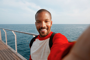 Handsome smiling young African-American hipster man taking selfie with happy face at the beach. Blue sea on the blurred background