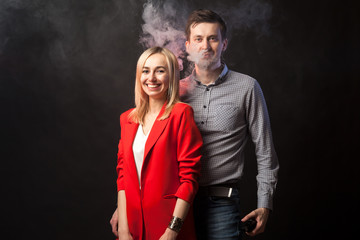 Young beautiful white blond girl in brightly red suit with jacket and white blouse and tall dark-haired guy hugging against smoke and isolated black background