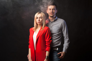 Young beautiful white blond girl in brightly red suit with jacket and white blouse and tall dark-haired guy hugging against smoke and isolated black background