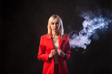 Young beautiful white blond girl in a bright red strict suit with a jacket and white blouse looks at the camera on a background of colored blue and pink smoke on a black isolated background