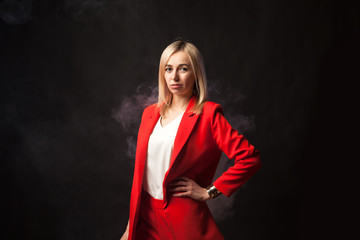 Fototapeta premium Young beautiful white blond girl in a bright red strict suit with a jacket and white blouse looks at the camera on a background of colored blue and pink smoke on a black isolated background