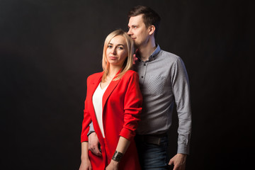 Young beautiful white blond girl in brightly red suit with jacket and white blouse and tall dark-haired guy hugging and kissing on isolated black background