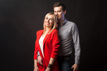 Young beautiful white blond girl in brightly red suit with jacket and white blouse and tall dark-haired guy hugging and kissing on isolated black background