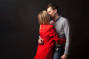 Young beautiful white blond girl in brightly red suit with jacket and white blouse and tall dark-haired guy hugging and kissing on isolated black background
