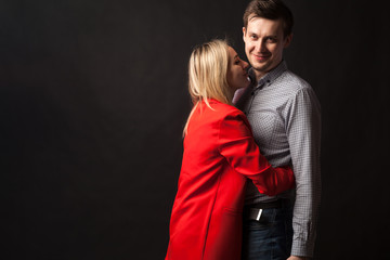 Young beautiful white blond girl in brightly red suit with jacket and white blouse and tall dark-haired guy hugging and kissing on isolated black background