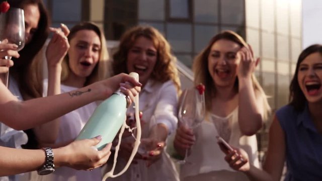 Beautiful young ladies celebrate. Hen-party. Shaking a bizarre bottle of champagne and letting it spraying. The girl pours a drink into glasses. Outside. Glamorous party. Slow motion