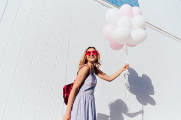 Beautiful funny curly woman in pink sunglasses enjoying the sunny day, holding air balloons, looking top, cheerfully smiling, wearing summer dress, with backpack. Outside.