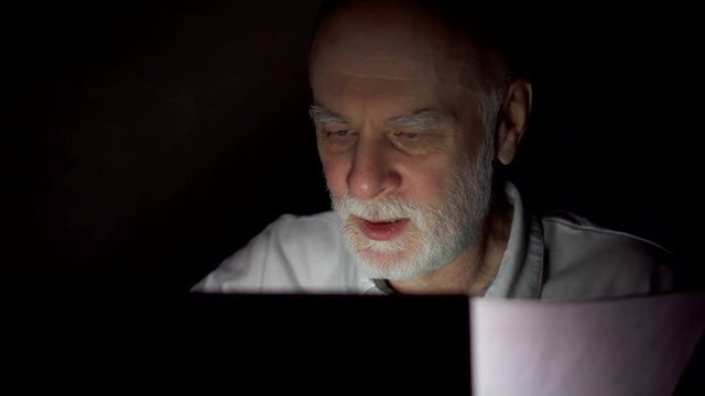 Tired overworked businessman at home working on laptop with documents late at night. Checking data for annual financial report. Secretly using computer hiding from people. Dark only face illuminated