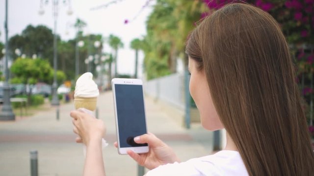 Young woman in white t-shirt taking photo of ice cream cone outdoors in summer. Happy female teenager photographing vanilla ice cream on her camera phone on street in slow motion