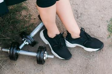 Closeup image of a woman legs in black sneakers near dumbbells outdoor on ground