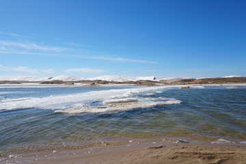 Beaurtiful sunny day along the lake shore on spring day with ice and snow in water, blue sky in the background, sandy beach in foreground